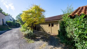 Un chemin de campagne menant à une maison avec un toit en tuiles, entourée de verdure et d’un arbre aux feuilles jaunes.
