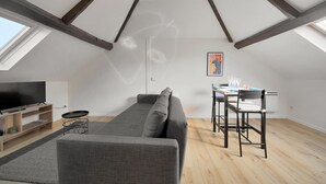Een modern loft appartement met hoge plafonds, zichtbare houten balken, een grijze bank, een eettafel met stoelen en een flatscreen televisie.