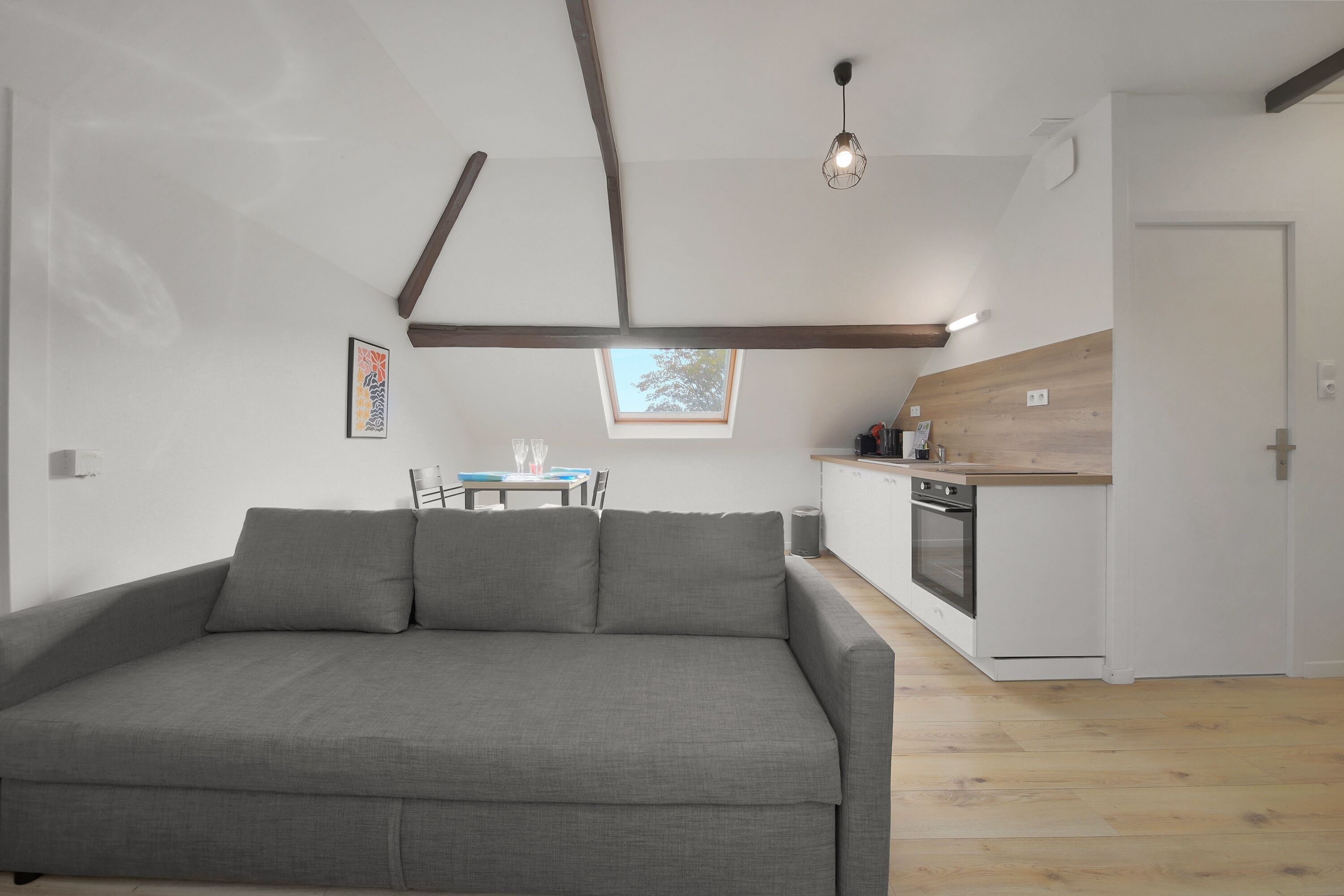 Una sala de estar moderna con un sofá gris, una mesa de comedor con sillas, una zona de cocina con armarios de madera y un tragaluz.
