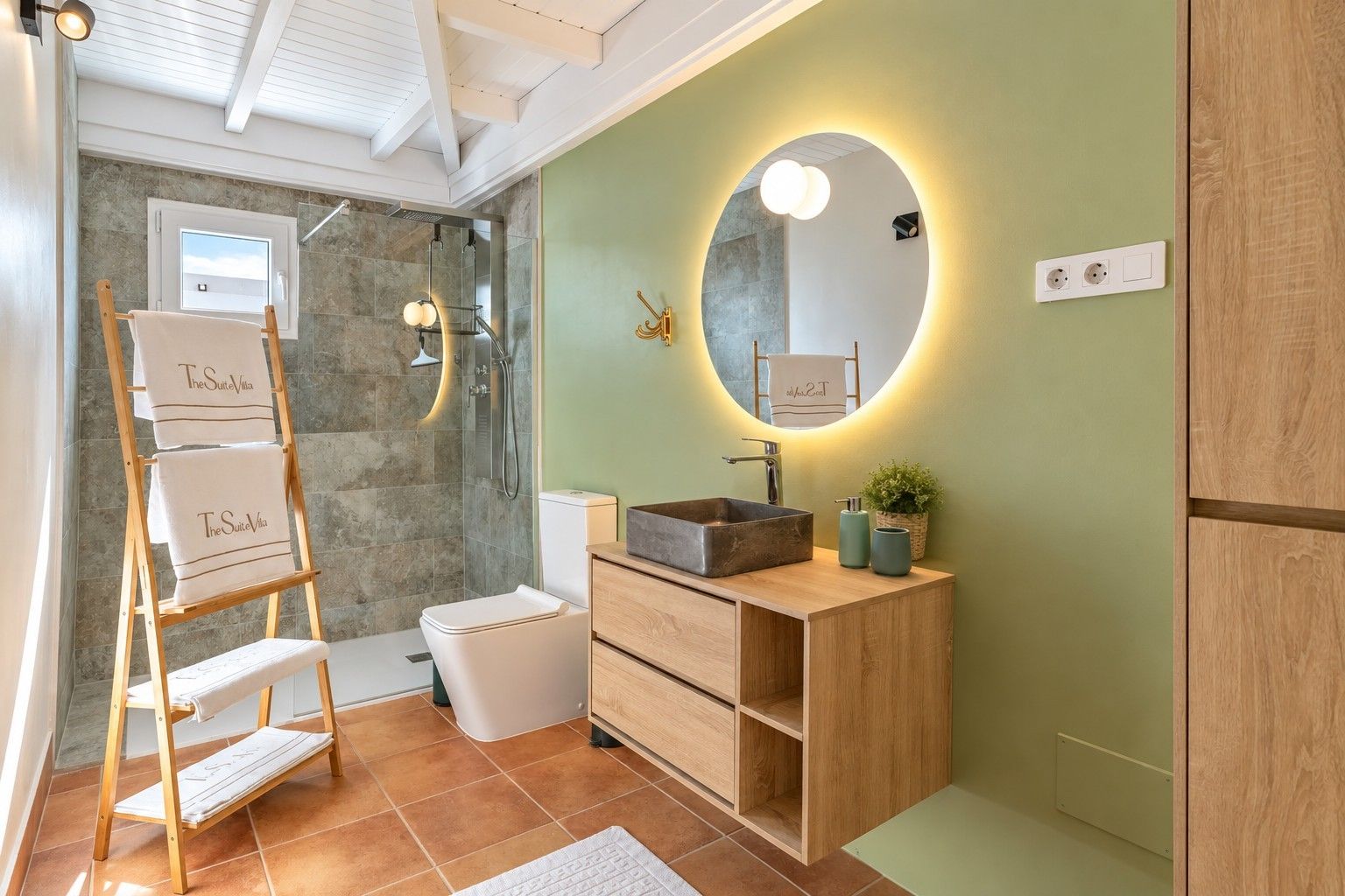 Un baño con tocador de madera, inodoro blanco, cabina de ducha de vidrio y un perchero con toallas de 'The Suite Villa'.