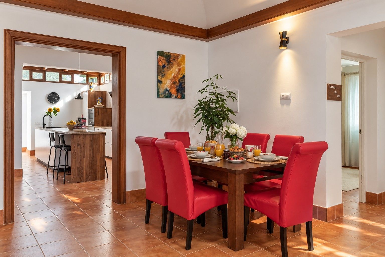 Un comedor con mesa de madera y sillas rojas, una cocina al fondo y un cuadro en la pared.