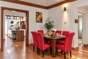 A dining area with a wooden table and red chairs, a kitchen in the background, and a painting on the wall.