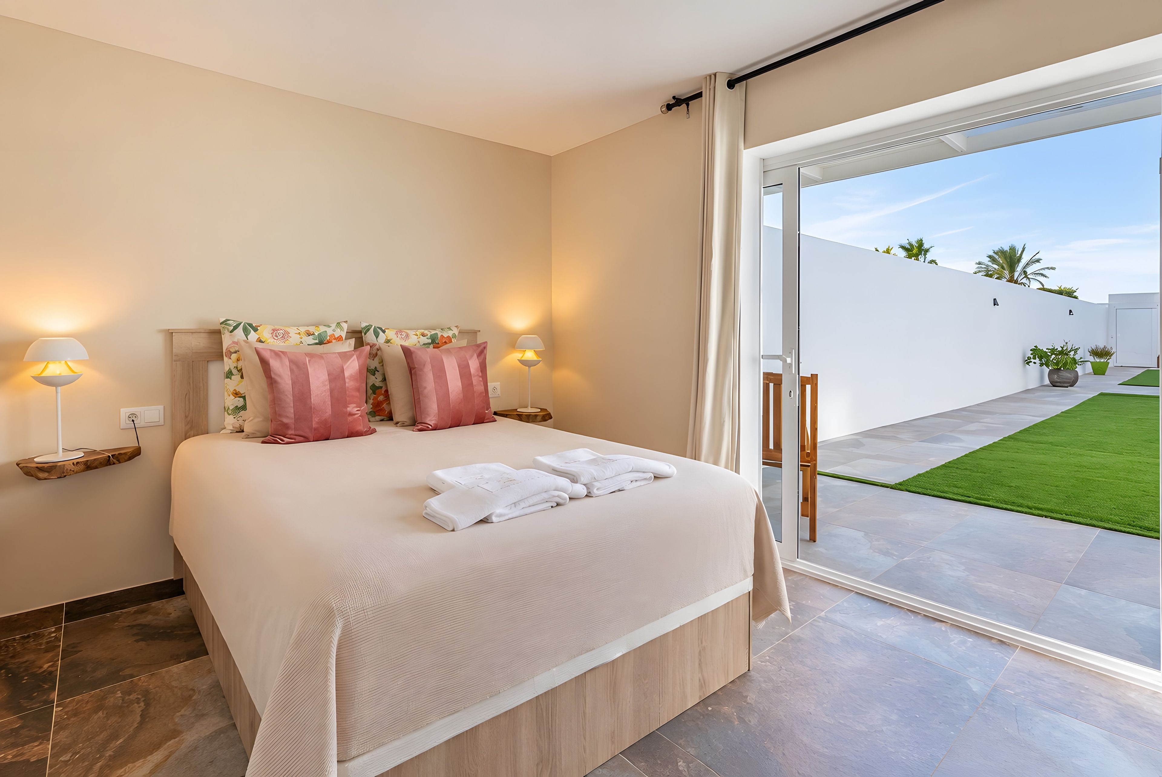 Ein modernes Schlafzimmer mit einem großen Bett, zwei Nachttischlampen und Blick auf eine grüne Rasenfläche durch eine offene Schiebetür.