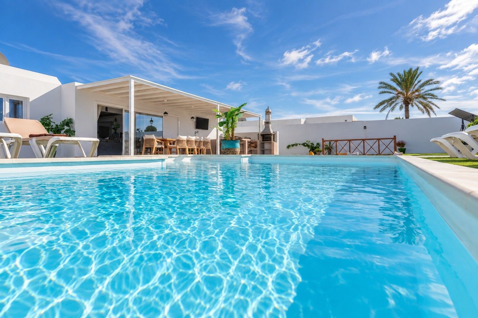 Piscina con sillas de descanso y una zona de estar techada, rodeada de paredes blancas y una palmera.