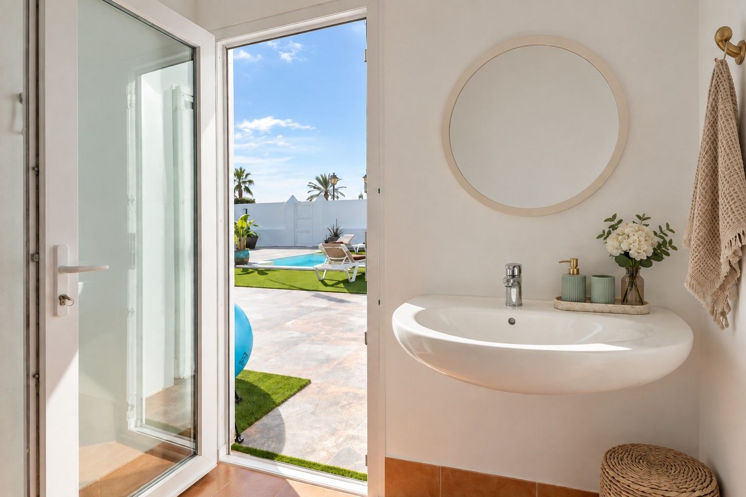 Un baño con un lavamanos blanco, un espejo redondo y vistas a una piscina y jardín.