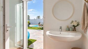 Un baño con un lavamanos blanco, un espejo redondo y vistas a una piscina y jardín.