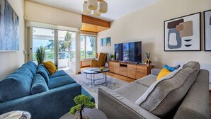 A modern living room with a sofa, coffee table, and TV. There is a sliding glass door leading to a balcony. - Berengaria Court - Sea Views 2bdr Apartment (Limassol)