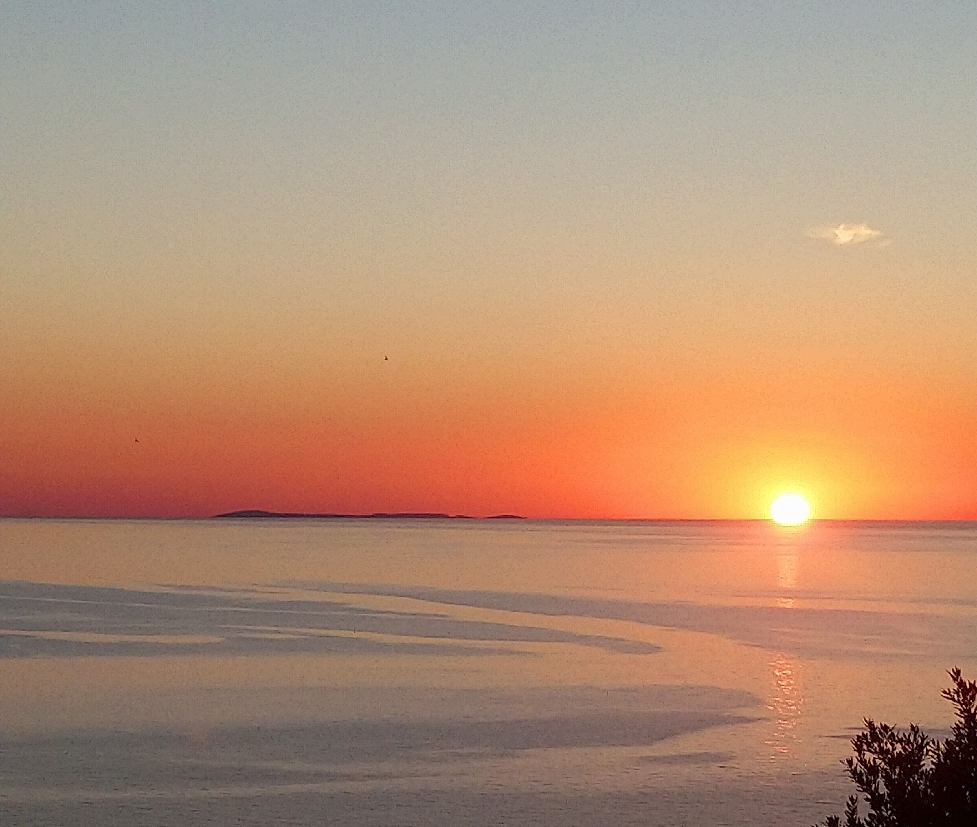 Un coucher de soleil sur une étendue d’eau calme, avec une masse terrestre visible au loin.