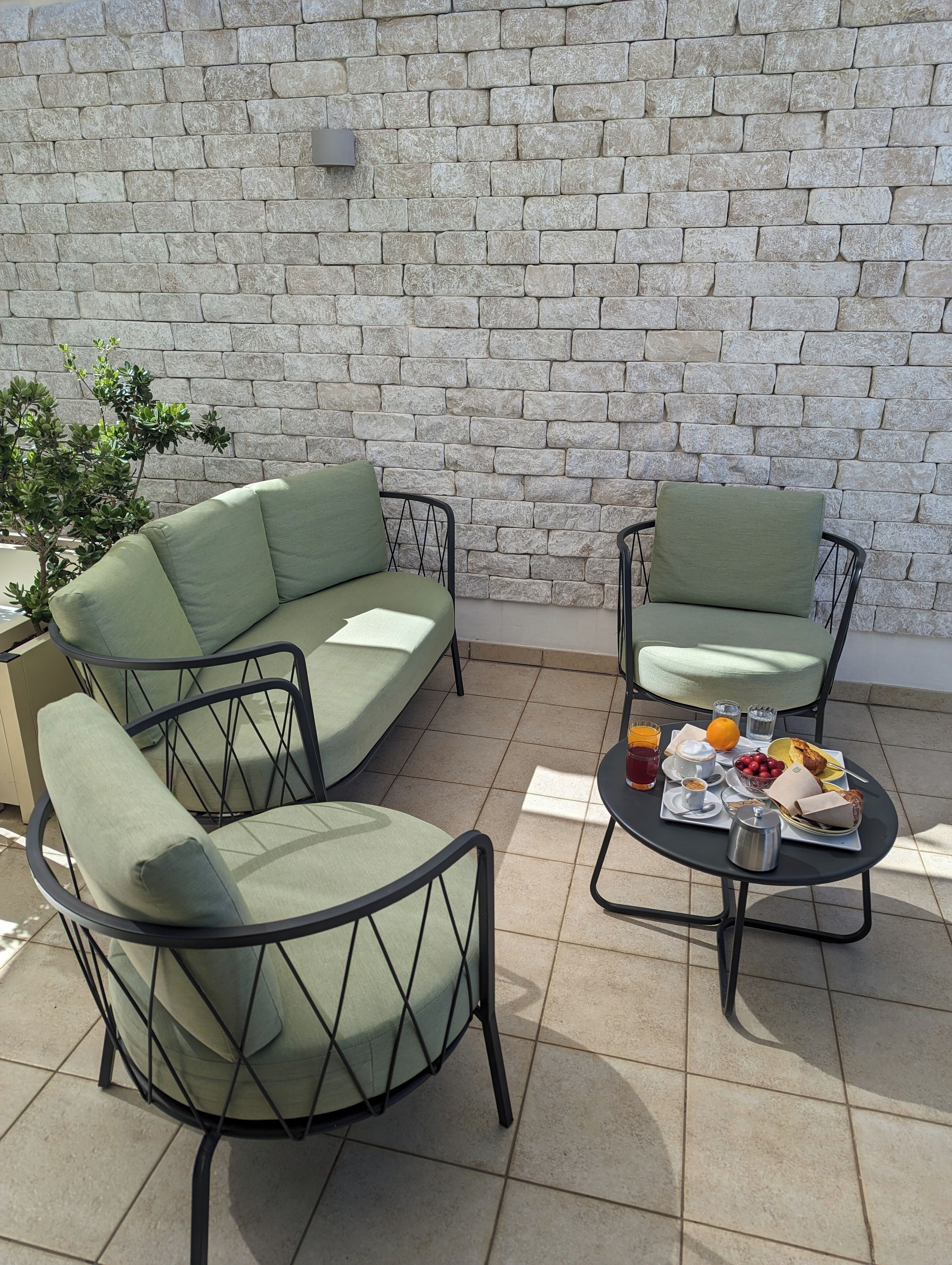 Un patio con muro de piedra, dos sillas acolchadas verdes, una pequeña mesa redonda con bebidas y aperitivos, y una planta en maceta.