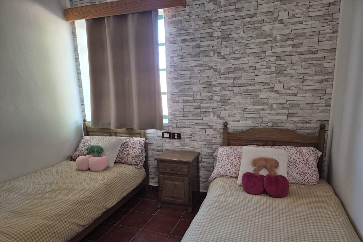A bedroom with two single beds, a nightstand, a wall-mounted power outlet, and a window with curtains.