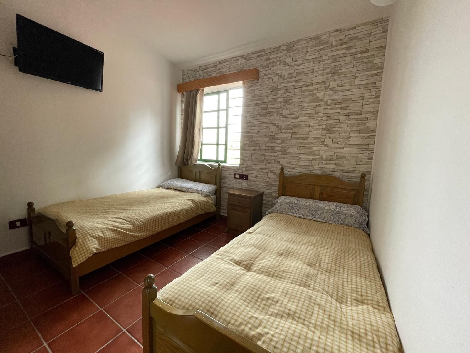Una habitación con dos camas de madera, una pared con diseño de piedra, un ventanal con cortinas y un televisor de pantalla plana montado.