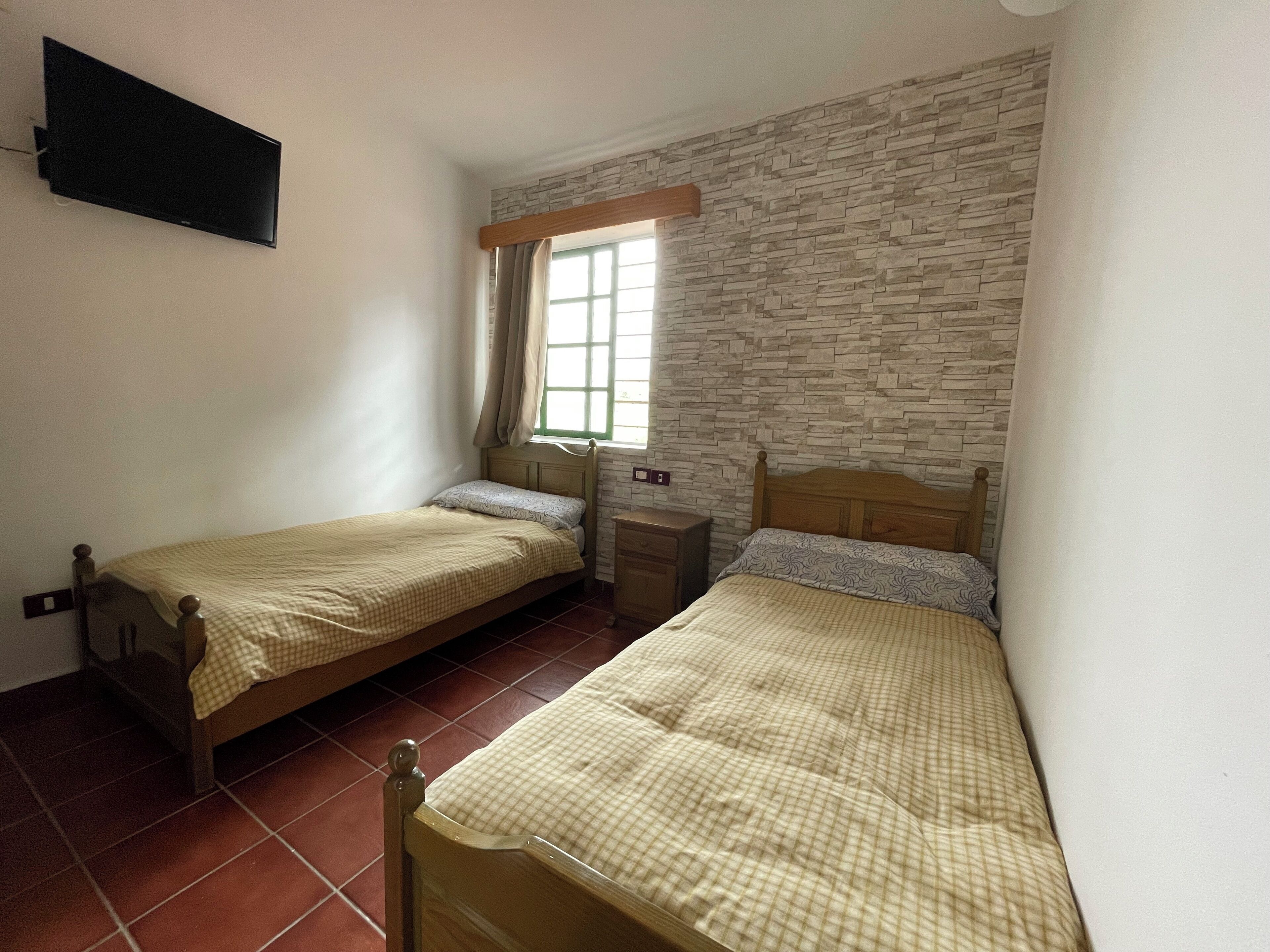 Une chambre avec deux lits simples, une table de chevet, un téléviseur fixé au mur et une fenêtre avec des rideaux.