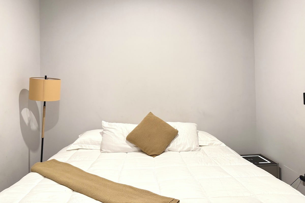 A neatly made bed with white bedding and a brown throw blanket, a floor lamp, and a small bedside table with a lamp.