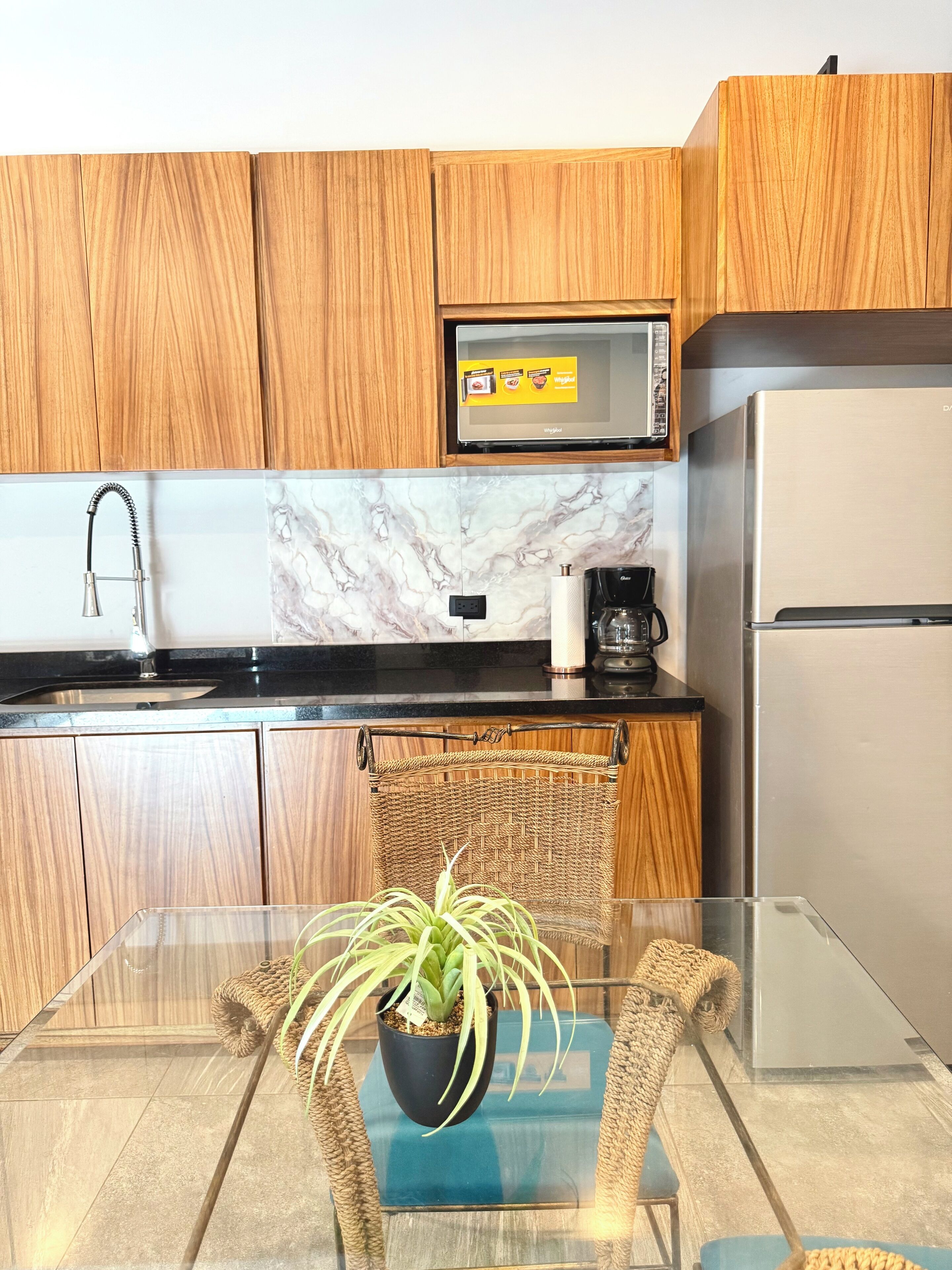 A modern kitchen with wooden cabinets, a stainless steel refrigerator, a microwave, and a glass table with a potted plant.