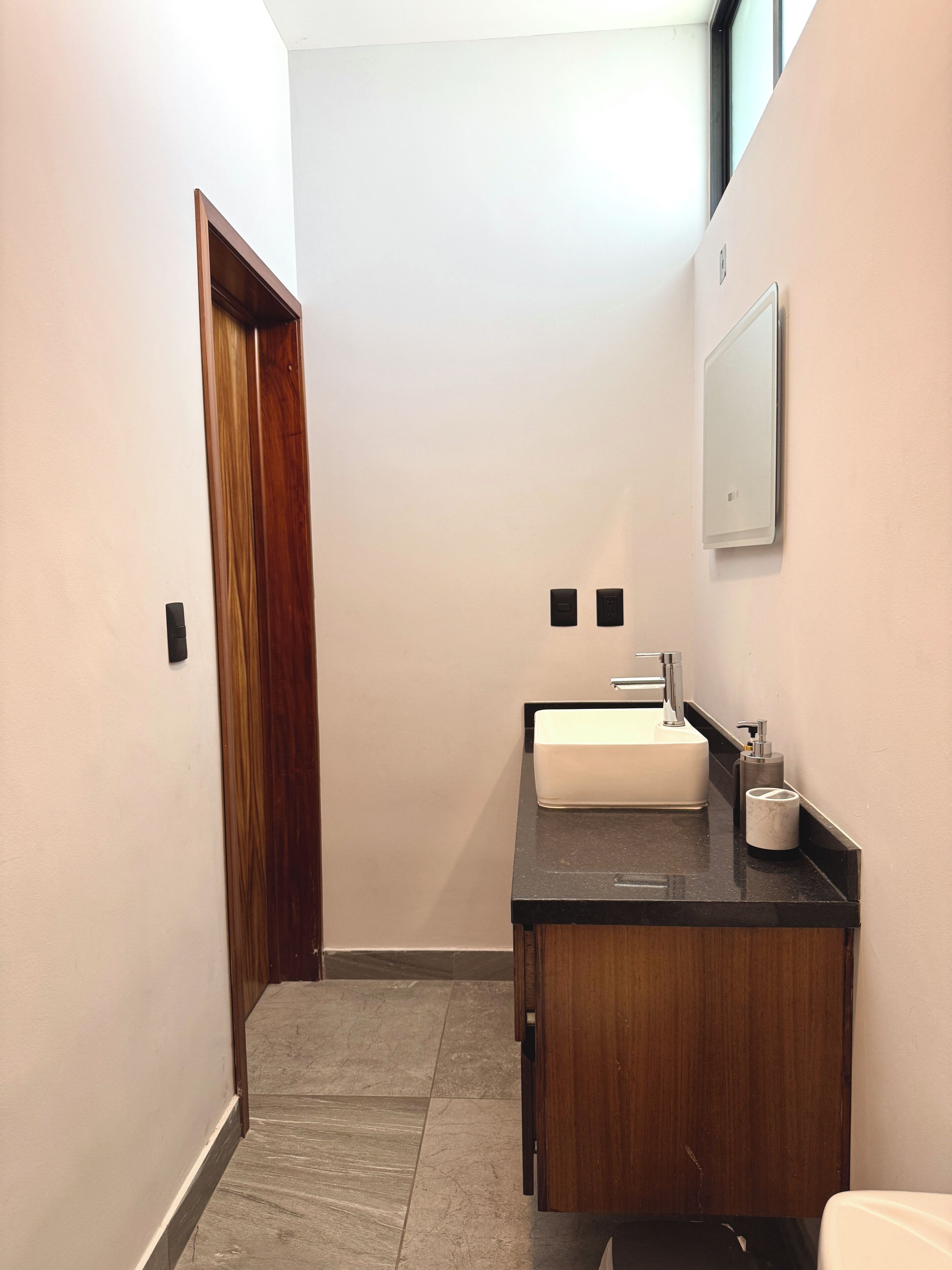 Un couloir étroit avec une porte en bois, un lavabo blanc avec un plan de travail noir et un vase en marbre.