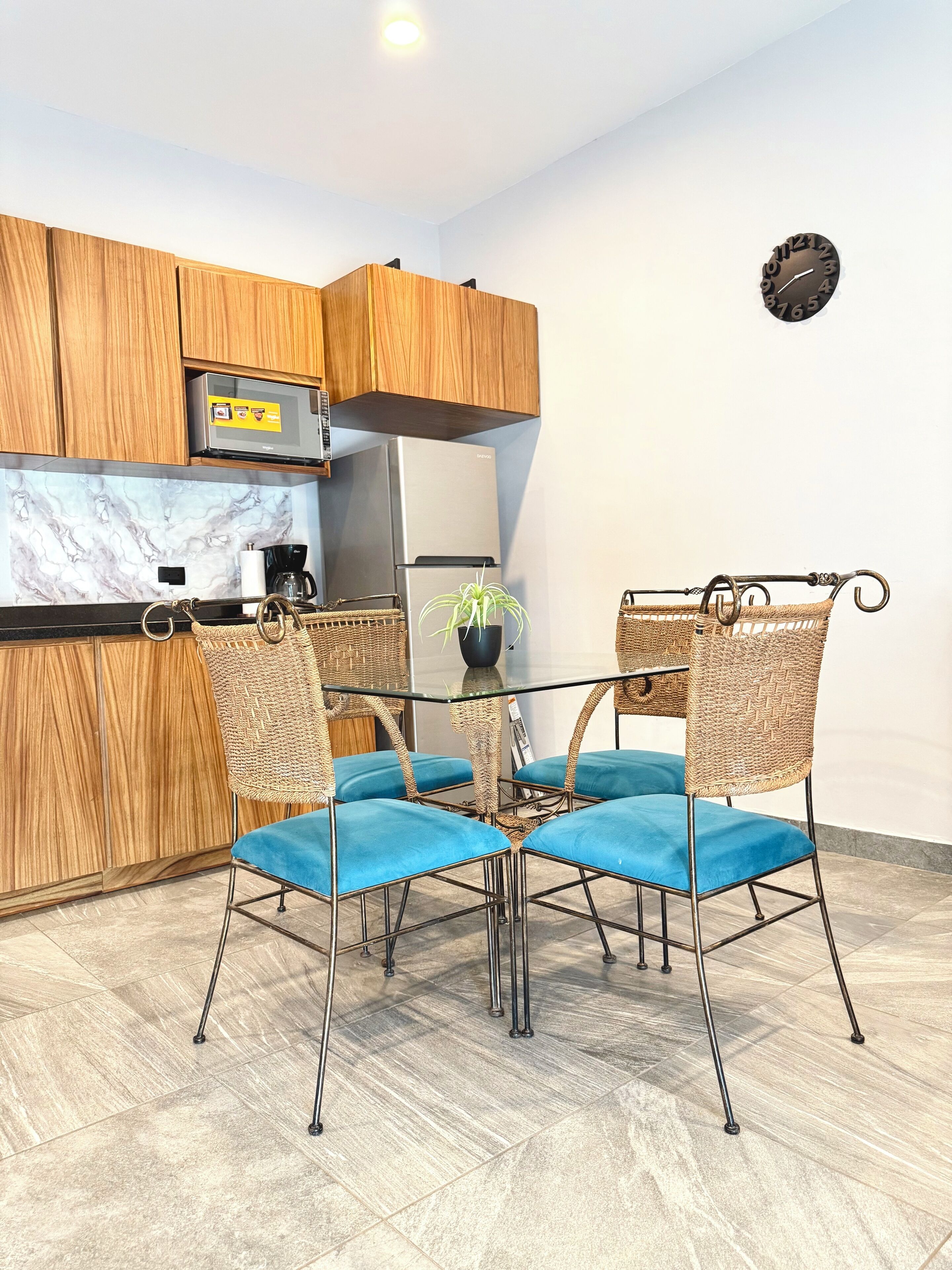 Une cuisine compacte dotée de meubles en bois, d’un réfrigérateur en acier inoxydable et d’une table à manger en verre, accompagnée de quatre chaises bleues à coussin.
