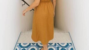 Eine Frau in einem senffarbenen Kleid steht in einer Diele mit weißen Wänden, einem Wandbild mit Fischen und gemusterten Bodenfliesen.