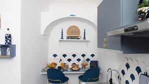 Eine kompakte Küche mit weiß-blauer Farbgestaltung, ausgestattet mit einem Esstisch und Stühlen, einer Hängelampe und einem kleinen Regal mit Dekoration.