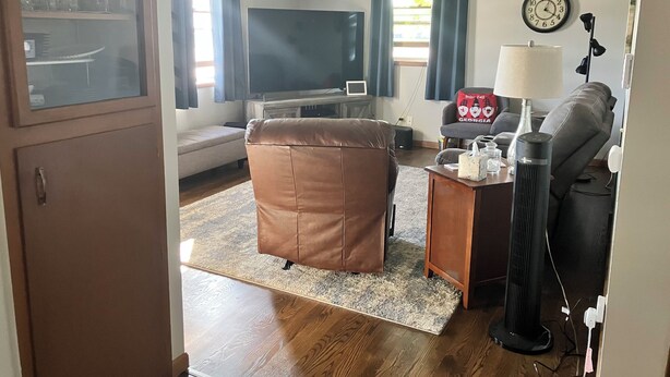A living room with a sofa, a brown leather ottoman, a coffee table, a TV, a lamp, and a clock on the wall.