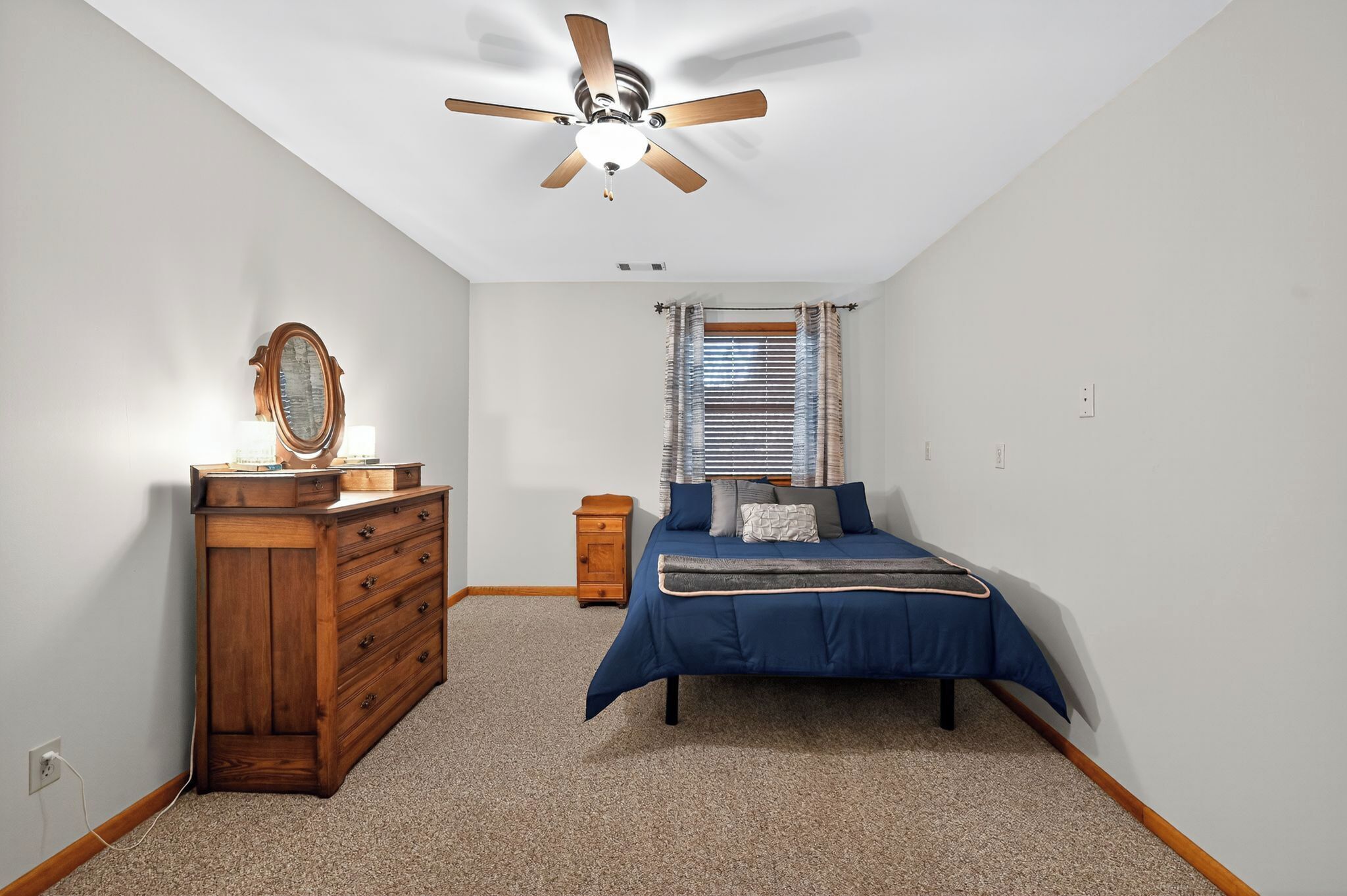 A bedroom with a wooden dresser, a bed with a blue bedspread, a ceiling fan, and a window with blinds.