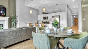 Un salon moderne avec un canapé gris, une table à manger entourée de chaises et, au fond, une cuisine.