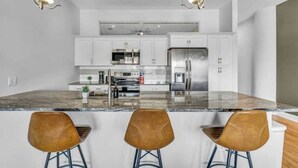 A modern kitchen with white cabinets, stainless steel appliances, and a marble countertop. There are three stools with metal legs. - The Halle Downtown 1BD / FREE Parking/Gym/Security (Cleveland)