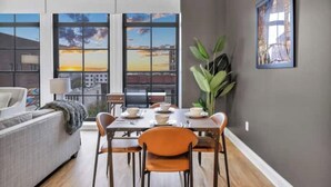Comedor con una mesa dispuesta para cuatro, ventanas amplias con vistas a la ciudad, un sofá gris y una planta en maceta.