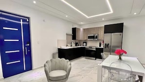 Une cuisine moderne avec une porte bleue, un coin repas avec une table et des chaises blanches, et un fauteuil gris.