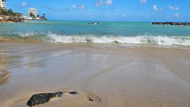 Une plage à l’eau turquoise et limpide, avec un rivage sablonneux et des affleurements rocheux.