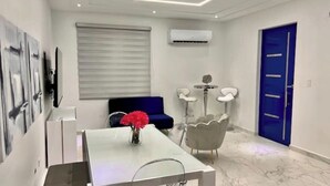 Une chambre d’hôtel moderne avec un plafond blanc, une porte bleue, un canapé gris, une table blanche ornée d’un vase de fleurs et un appareil de climatisation blanc.