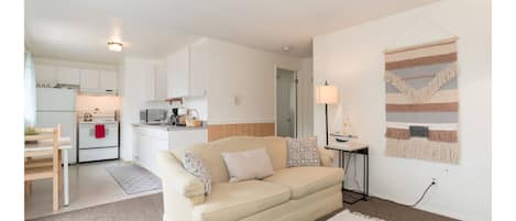 A living room with a beige sofa, a small table, and a lamp. There is a kitchen in the background with white cabinets and a microwave.