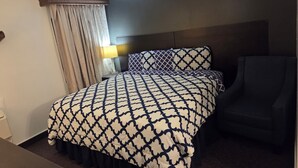 Quarto de hotel com cama, cadeira, mesa de cabeceira, luminária e janela com cortinas.