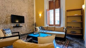 A living room with a stone wall, a ceiling fan, a TV, a sofa, a coffee table, and a bookshelf. - Casa Axuul - Colonial Property in Central Merida (Merida)