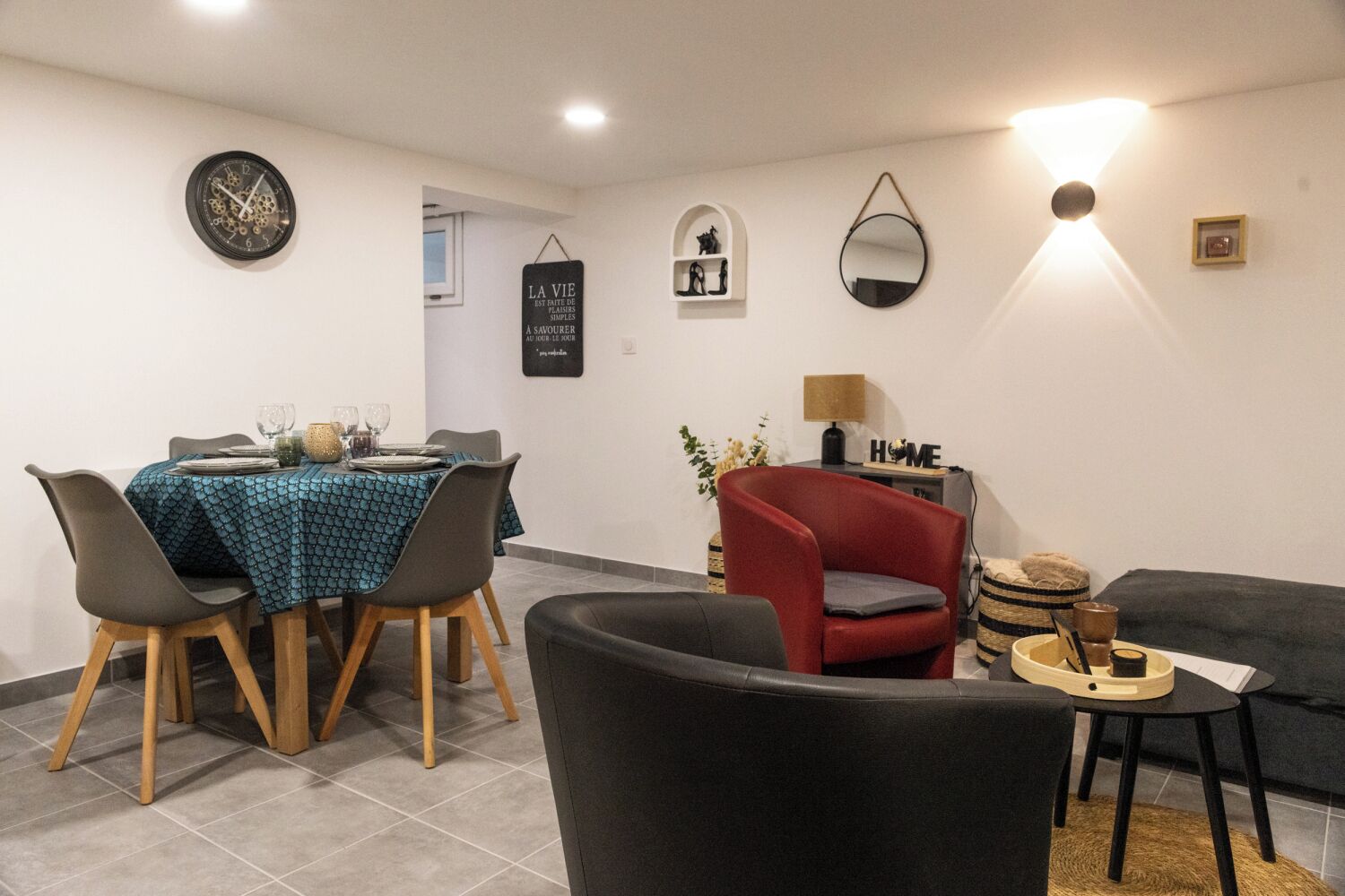Un comedor con una mesa dispuesta para cuatro, un sillón rojo, un sofá negro y una pequeña mesita de noche con una lámpara.