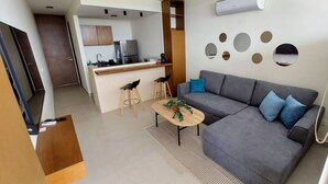 A modern living room with a grey sofa, a small coffee table, and a kitchen area with a fridge and bar stools. - Brisa de Montebello - By Kanaa (Mérida)
