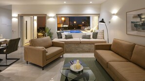 Ein modernes Wohnzimmer mit einem Glastisch, zwei Sofas, einem Essbereich und Blick auf einen Pool in der Nacht.