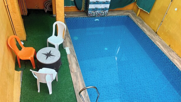 A small indoor pool with a blue tiled floor, surrounded by a yellow wall. There are two white plastic chairs and an orange plastic chair. - Serenity Bay Home Stay Pondicherry (Pondicherry)