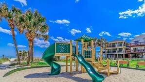 Een speeltuin aan het strand met een glijbaan, palmbomen en een gebouw op de achtergrond.