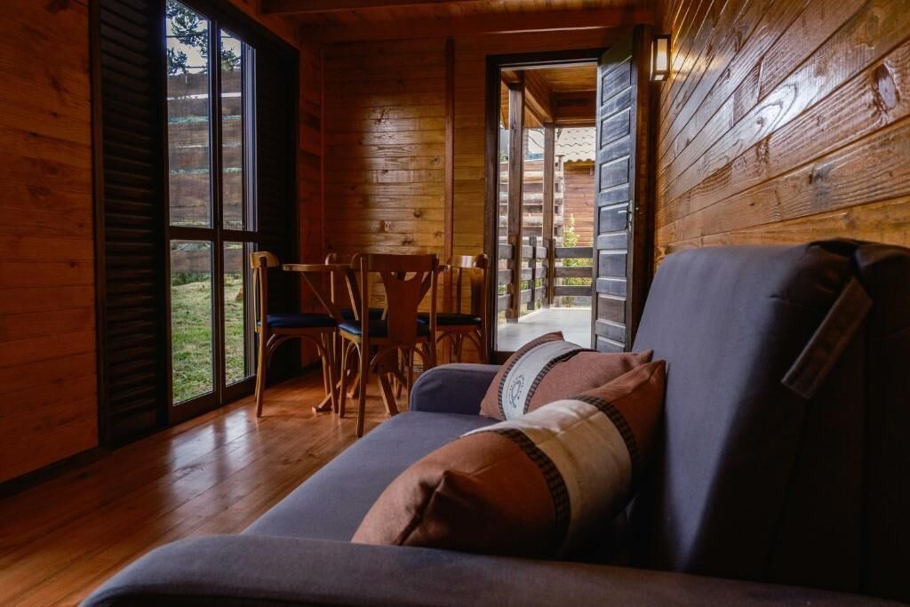 A cozy cabin interior with a wooden floor, a sofa with cushions, a dining table with chairs, and large windows.