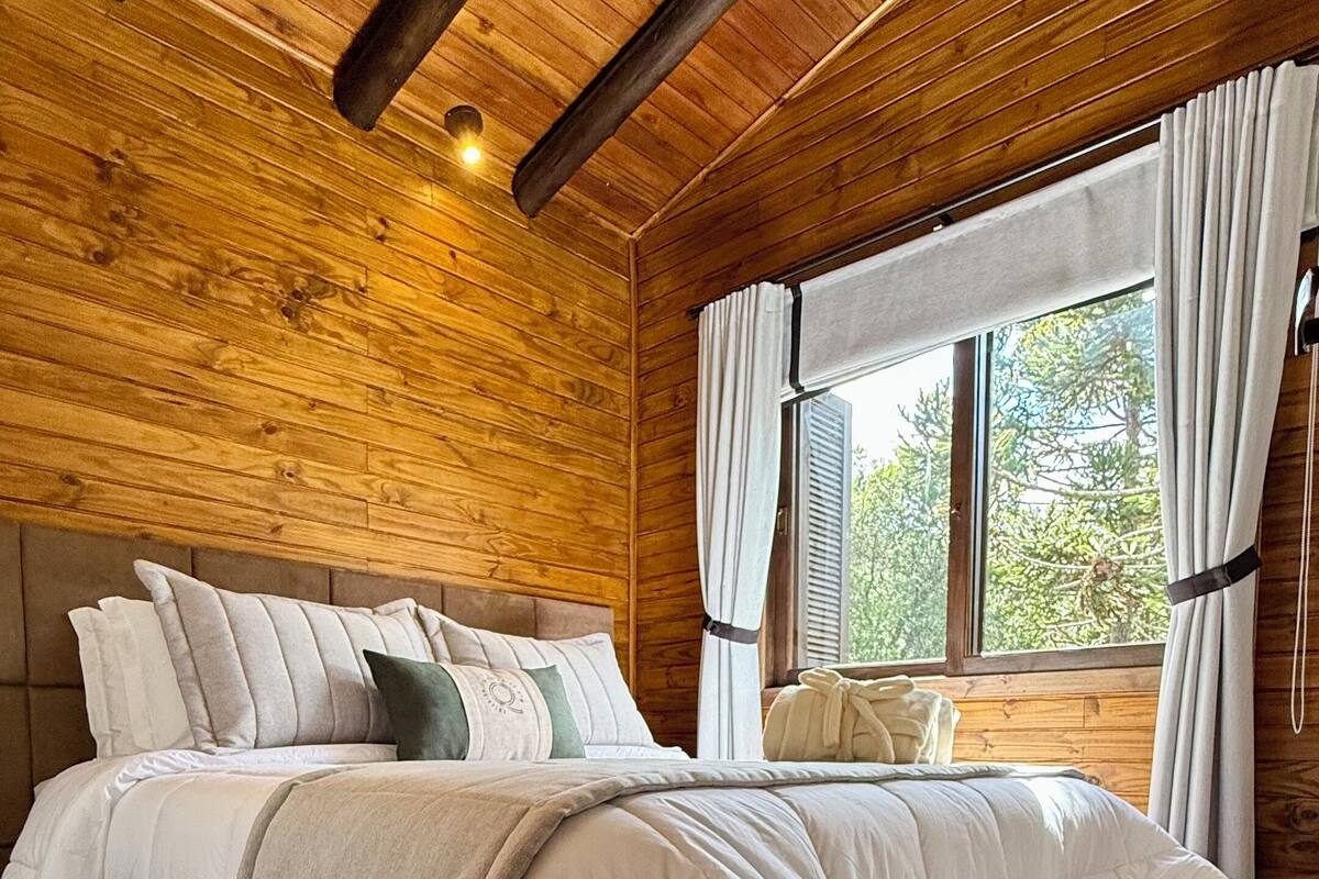 A wooden-paneled bedroom with a large bed, a window with curtains, and a pair of slippers on the floor.