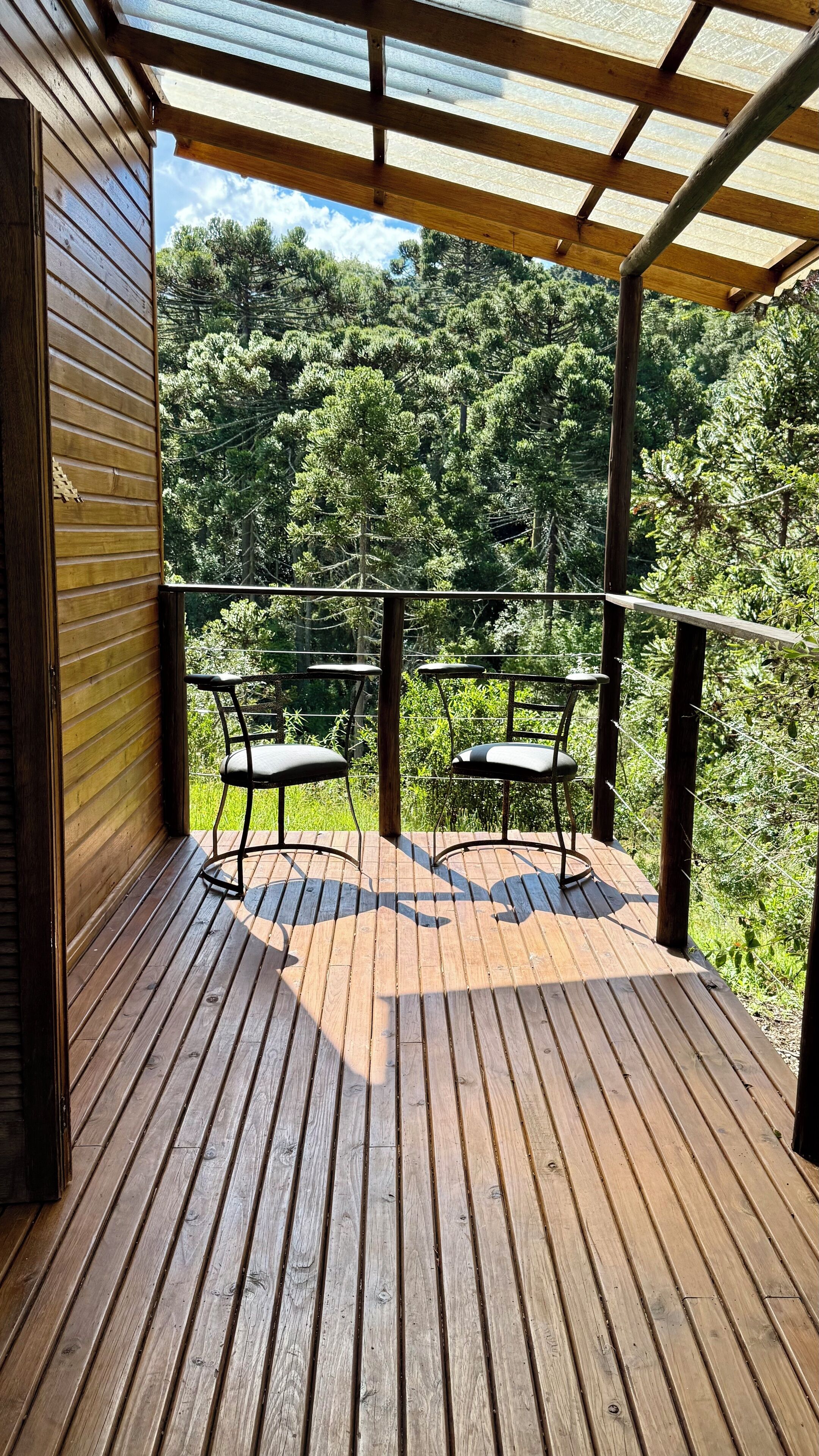 Une terrasse en bois avec deux chaises en métal et une balustrade en verre donnant sur une zone boisée.