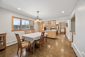 A dining area with a table and chairs, a kitchen with cabinets and a refrigerator, and a view through a window.