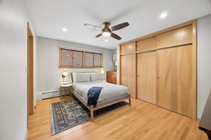 A bedroom with a bed, a ceiling fan, a window with blinds, a wooden wardrobe, and a nightstand with a lamp.