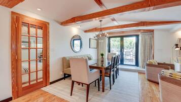 A spacious living area with a wooden ceiling, a dining table with chairs, a sofa, and a large window with curtains.