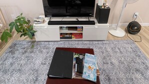 Un salon avec un téléviseur, une petite table avec des livres, une étagère décorative avec des figurines, un ventilateur sur pied et une plante en pot.