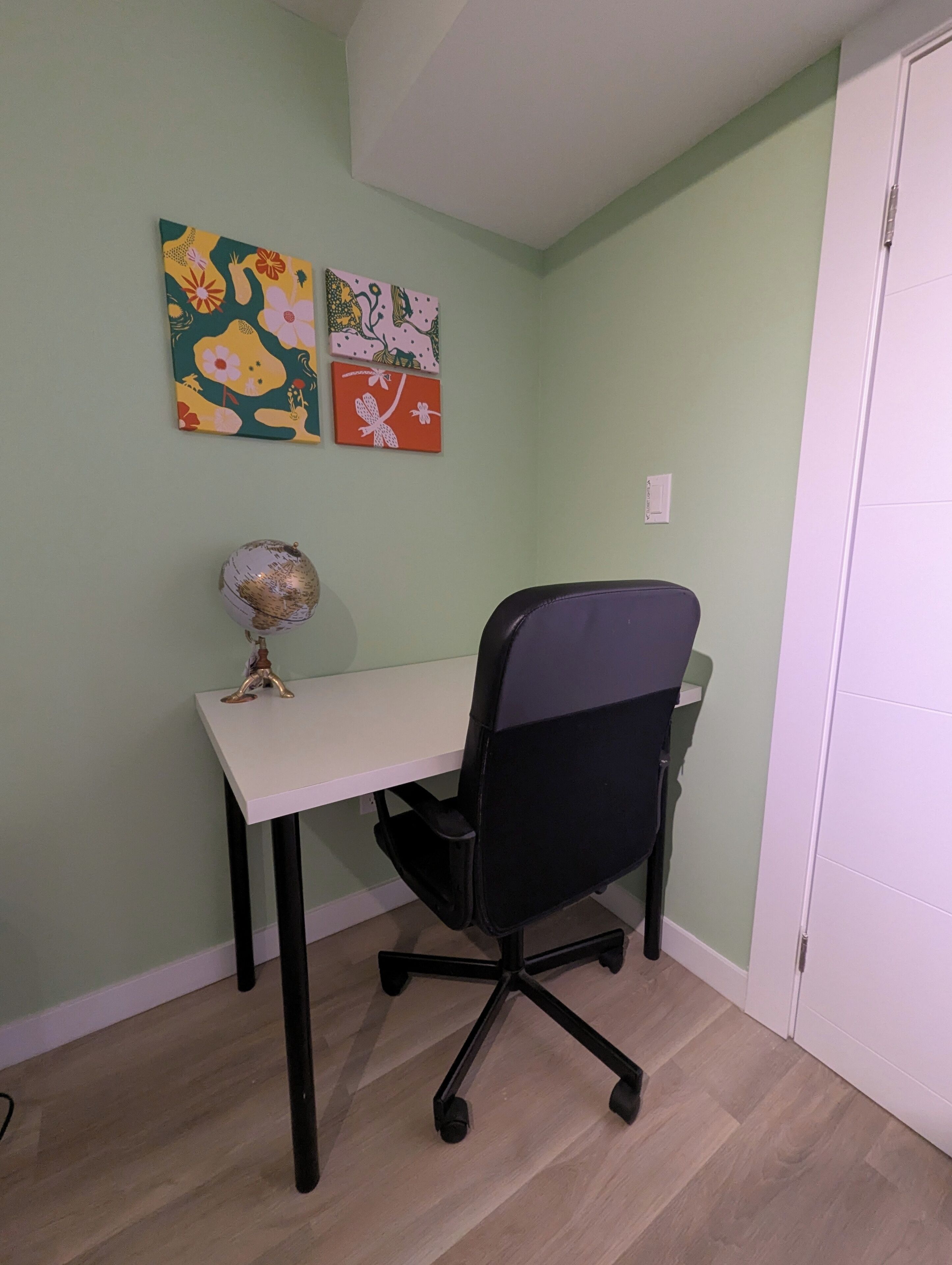 Una habitación con un escritorio blanco, una silla de oficina negra y un globo terráqueo sobre el escritorio. Hay tres cuadros coloridos en la pared.