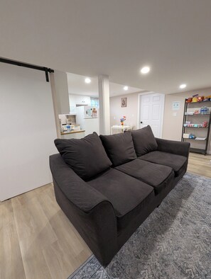 A modern living room with a grey sofa, a patterned rug, and a kitchen in the background. - Cozy & Spacious Family Friendly Basement Unit with Private Room and Bathroom (Westphal)