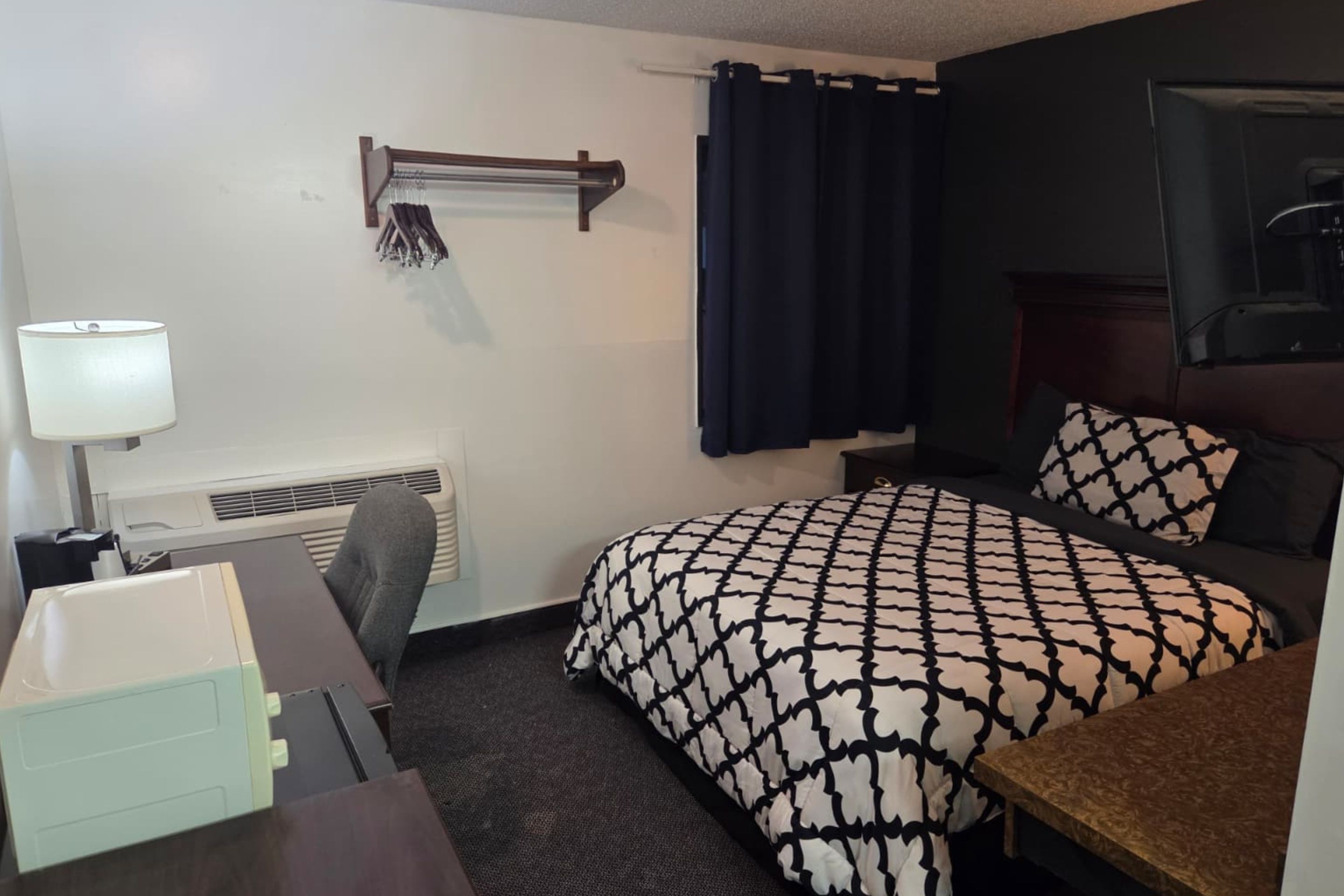 A hotel room with a bed, a desk, a chair, a lamp, a heater, and a curtain.