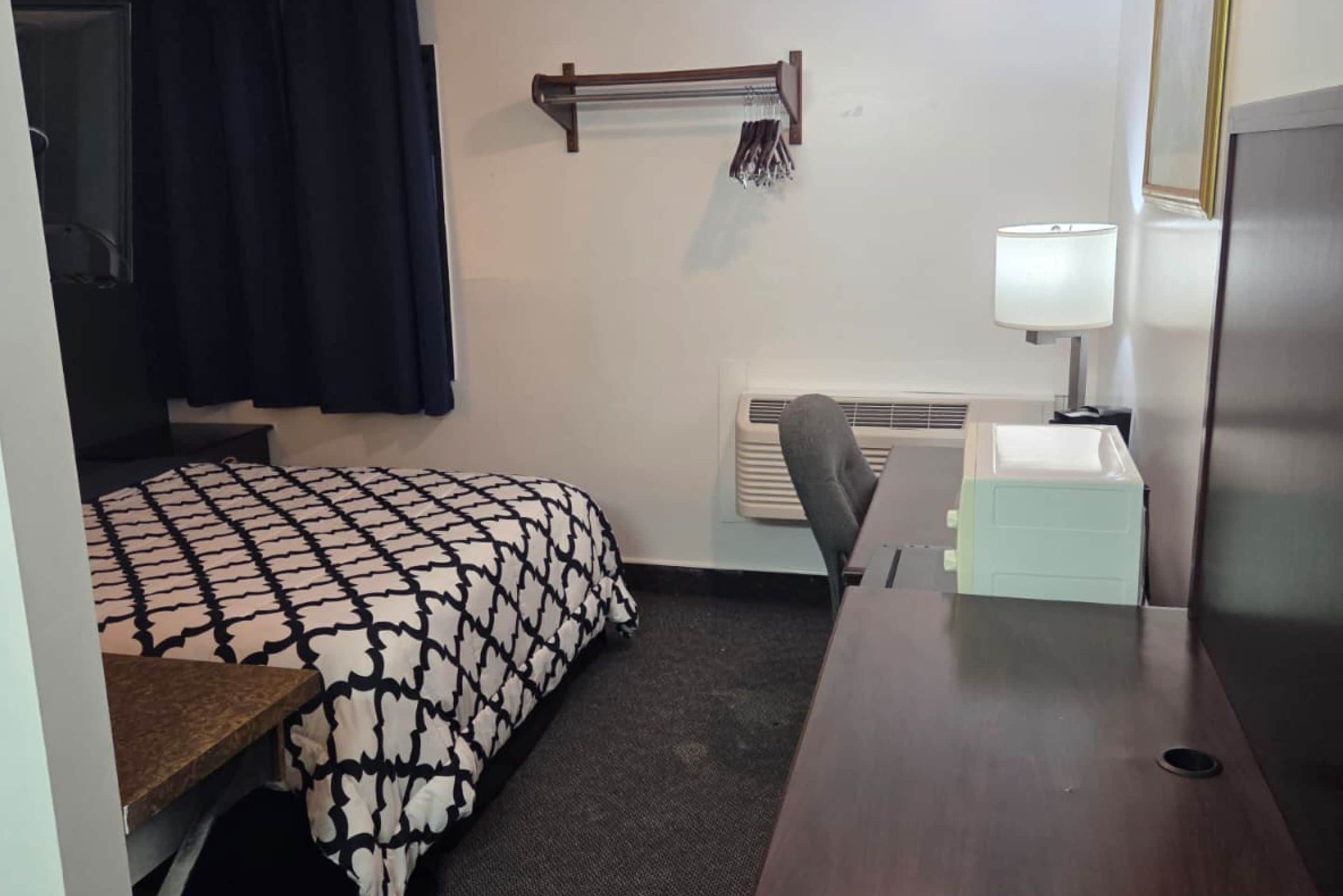 Habitación de hotel pequeña con cama, escritorio, silla, lámpara y radiador.