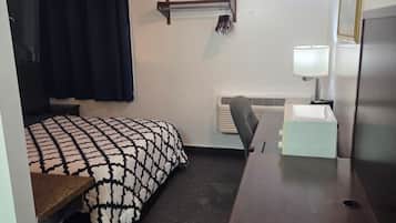 A compact hotel room with a bed, a desk, a chair, a lamp, and a radiator.
